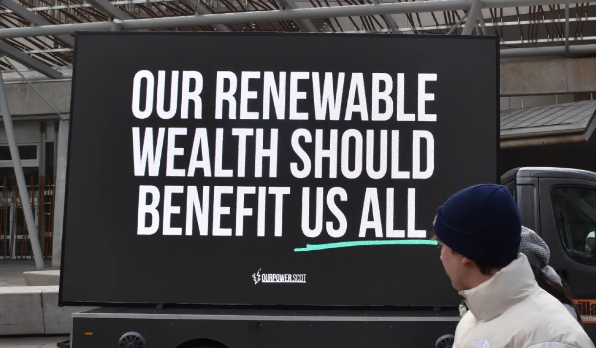 A person looking at a sign that says: Renewable wealth should benefit us all - Profit-hungry firms identified as top concern in renewable transition, as new campaign pushes for Scotland to get a fair share of energy wealth