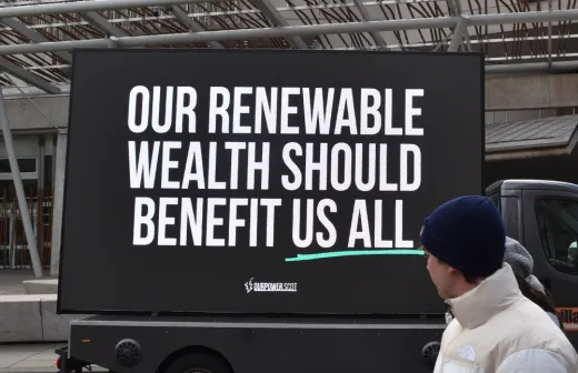 A person looking at a sign that says: Renewable wealth should benefit us all - Profit-hungry firms identified as top concern in renewable transition, as new campaign pushes for Scotland to get a fair share of energy wealth