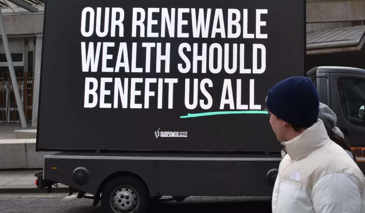 A person looking at a sign that says: Renewable wealth should benefit us all - Profit-hungry firms identified as top concern in renewable transition, as new campaign pushes for Scotland to get a fair share of energy wealth