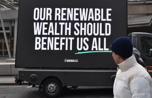 A person looking at a sign that says: Renewable wealth should benefit us all - Profit-hungry firms identified as top concern in renewable transition, as new campaign pushes for Scotland to get a fair share of energy wealth