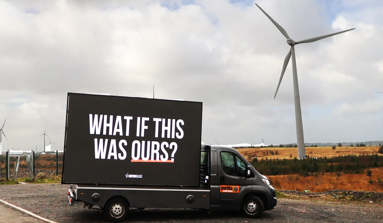 A van with a sign that says "What if this was our" in front of a wind turbine